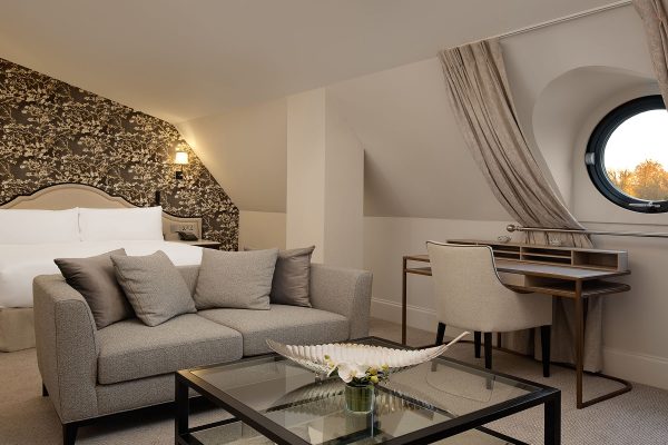 Attic guest room at Fairmont Windsor Park with plush sofa, floral wallpaper, and circular window view.
