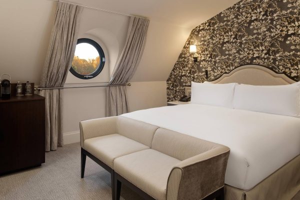 Loft-style hotel bedroom at Fairmont Windsor Park with round window, floral wallpaper, and neutral furnishings.