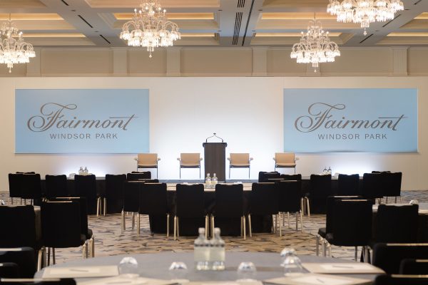 Conference room at Fairmont Windsor Park with chandeliers, podium stage, and screens displaying the hotel’s branding.