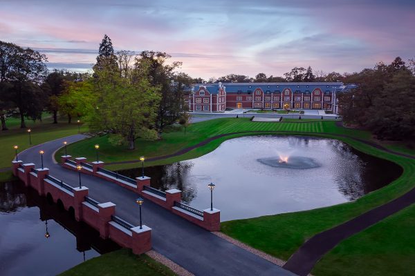 Stunning aerial view of Fairmont Windsor Park with a pond, a red brick bridge, and manicured lawns at sunset.