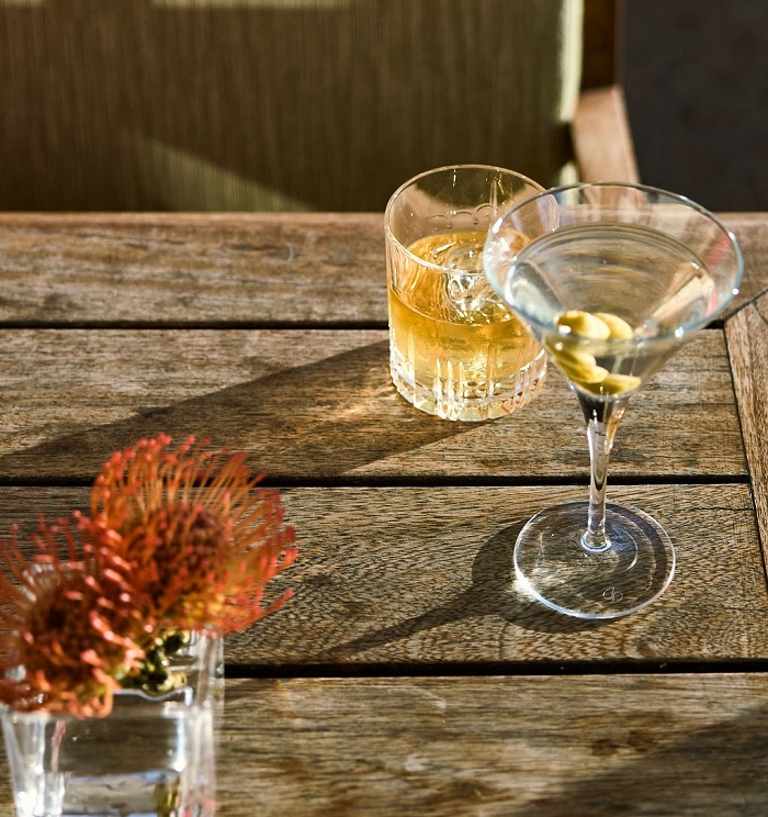3F6A5231 A whisky on the rocks and an olive martini on a sunlit wooden outdoor table, set with burnt orange flowers at Bascule Bar, Cape Grace.