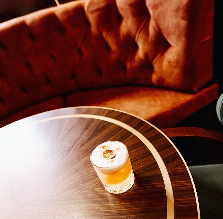 BASCULE-26 A frothy cocktail garnished with a dried orange slice on the round wooden table of Bascule Bar's velvet lounge seating at Cape Grace.