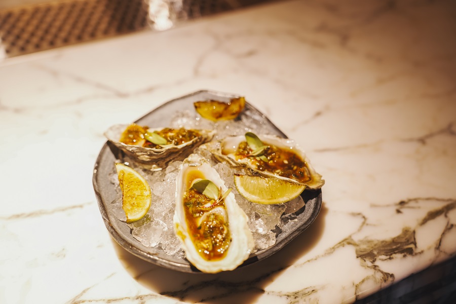 DSC02853 Three fresh oysters on the half shell, served on crushed ice with a savoury dressing and micro herbs, garnished with charred lemon wedges on a stone plate.