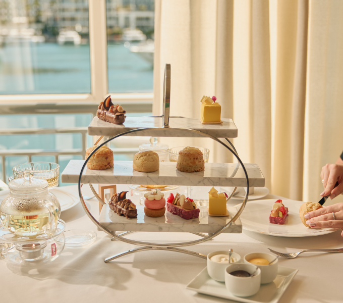 A guest savouring the three-tiered marble afternoon tea stand of buttermilk scones, gianduja chocolate cake and intricate petit fours, beside a glass teapot overlooking the V&A Waterfront marina at Cape Grace hotel.