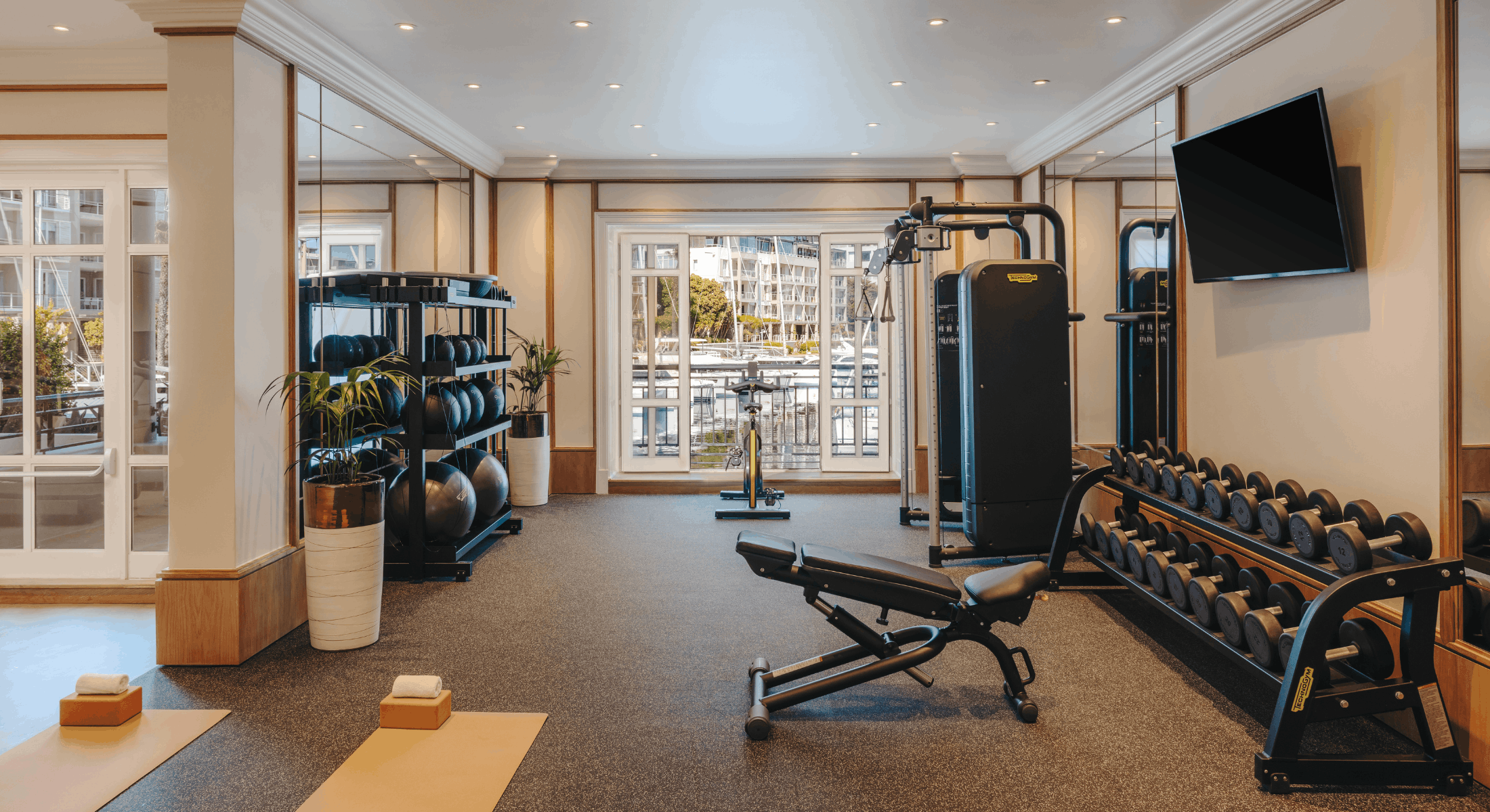 Bright, modern Fairmont Fit gym with dumbbells, weight machine, exercise bench, and yoga mats, overlooking the V&A Waterfront through large windows.