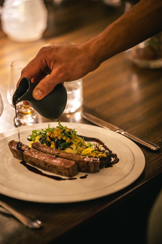 An Heirloom server finishing off a signature meat and vegetable dish tableside with a rich glaze at Heirloom Restaurant, Cape Grace.