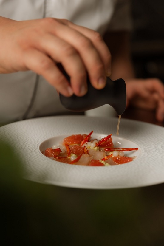 Chef adding a finishing dressing to a delicate seafood dish, plated on a textured white plate, at Heirloom Restaurant, Cape Grace.